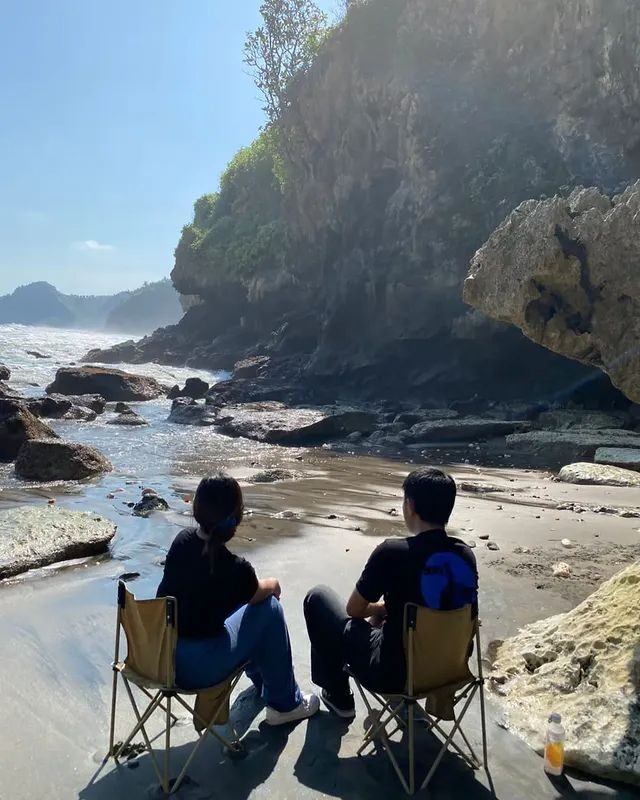 Dua orang duduk di kursi lipat menghadap laut di pantai berbatu bertebing