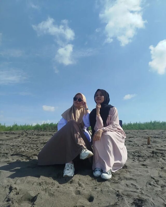 Dua orang duduk di kursi lipat di pantai berpasir gelap dengan langit biru