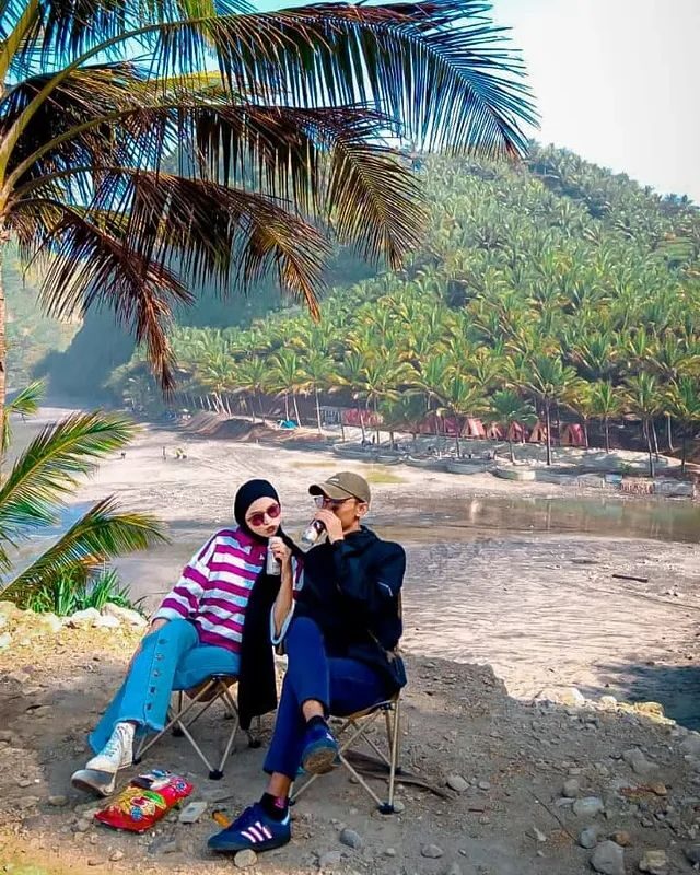 Dua orang duduk di kursi lipat di bawah pohon kelapa menghadap pantai
