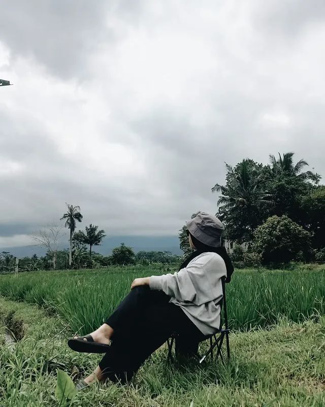 Seseorang duduk santai di kursi lipat menghadap hamparan sawah hijau