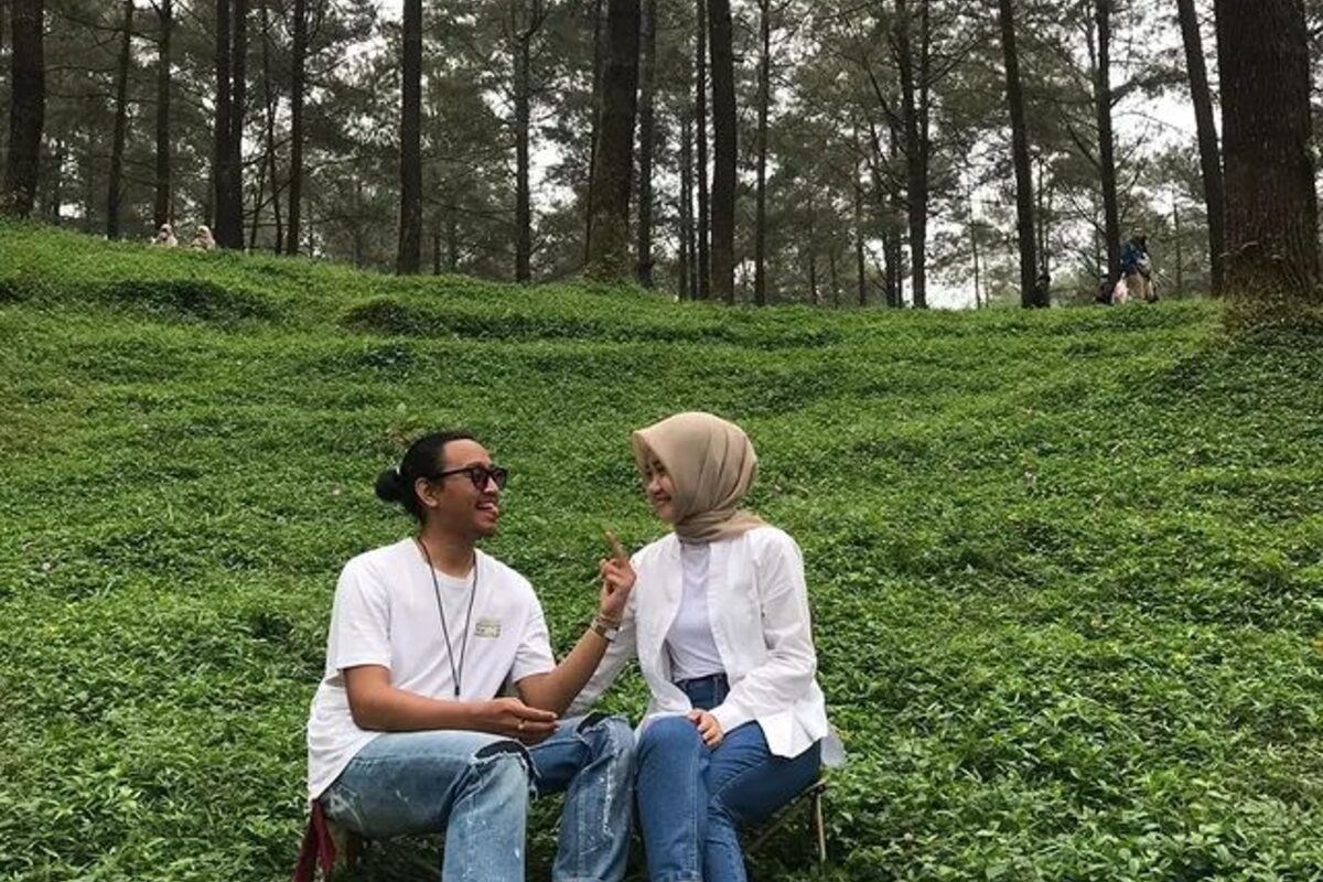 Dua orang duduk di kursi lipat di lereng berumput di antara pohon pinus, sedang berbincang