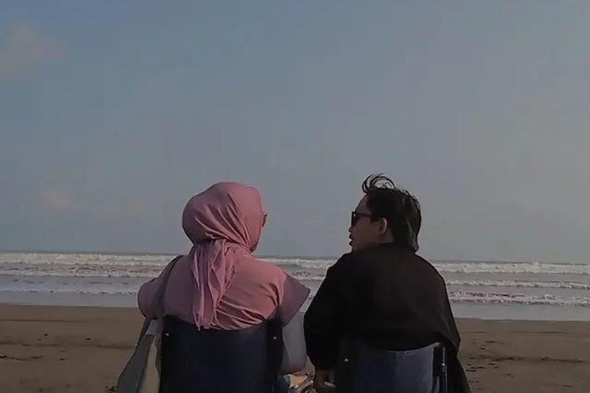 Dua orang duduk membelakangi kamera di kursi lipat di tepi pantai memandang laut